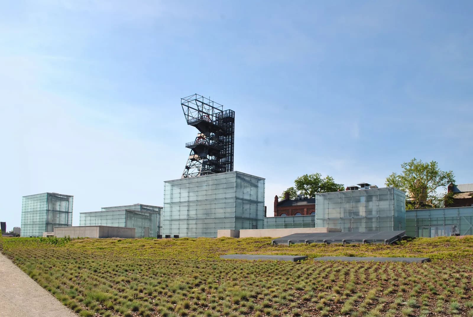 A modern building with glass block walls and an industrial tower behind it, contrasted with an expanse of green grass in the foreground under a clear sky.