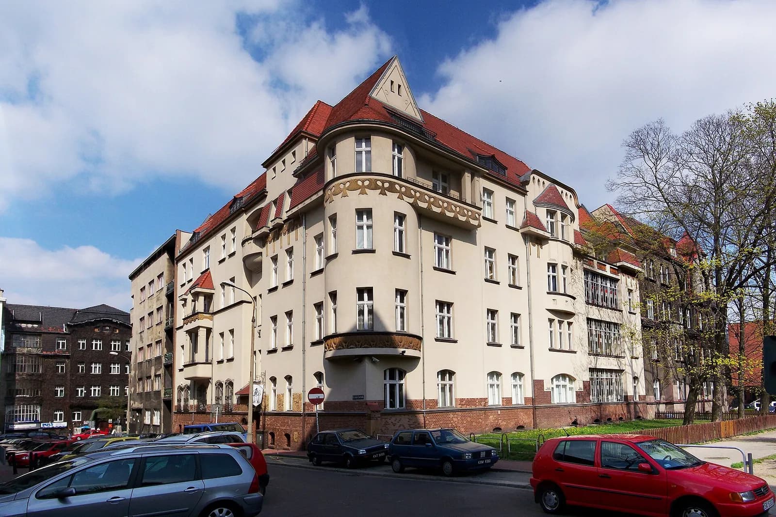 A large, multi-story building with a red-tiled roof and cream-colored walls, featuring many windows and intricate architectural details on a sunny day with scattered clouds, with cars parked along the street in the foreground.